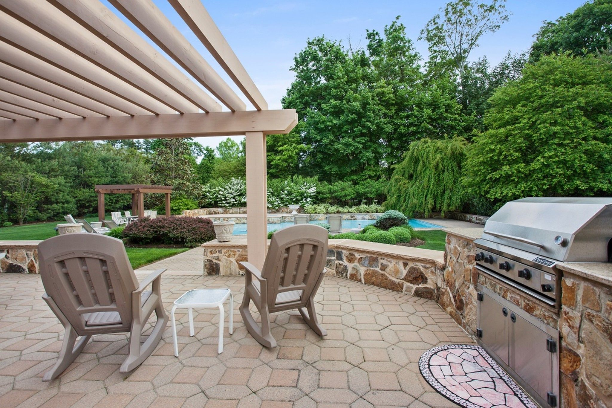 Backyard patio with a pergola, rocking chairs, a stone outdoor kitchen and a pool beyond
