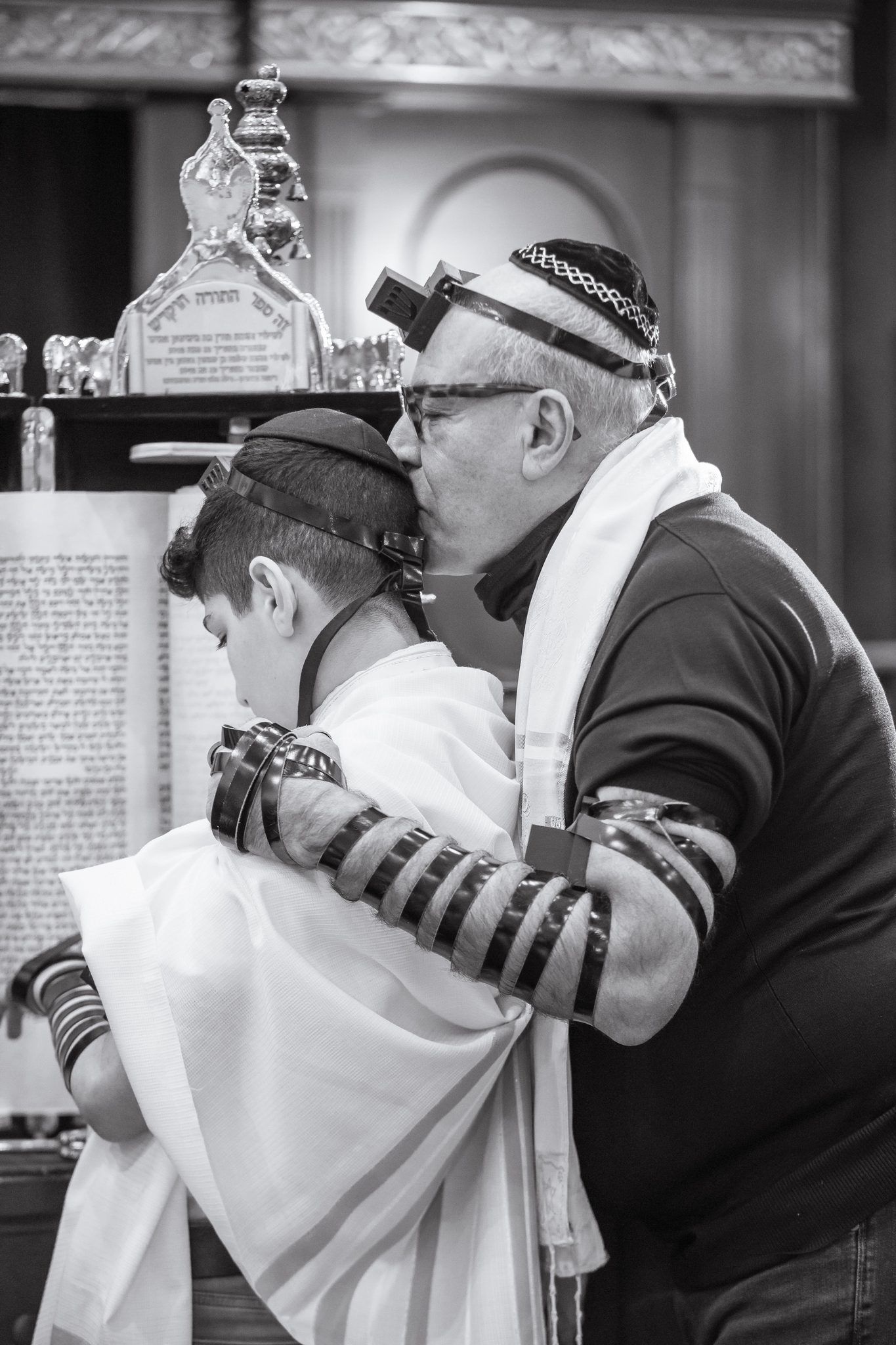 Black-and-white photo of a father kissing his bar mitzvah son's head at the Torah